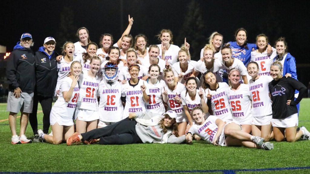 的Pomona-Pitzer women’s 长曲棍球 team gathers for a group photo on the field and raises their fingers in a number one gesture. 的 team wears white uniforms with Pomona Pitzer and their individual numbers on the front in blue and orange font.