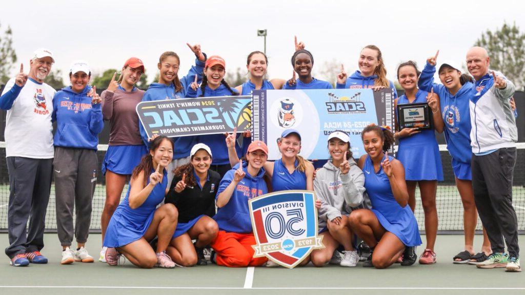 的Pomona-Pitzer women’s tennis team gathers on the court for a group photo. 的 team raises their fingers in a number one gesture and hold up signs for the SCIAC tournament championship.